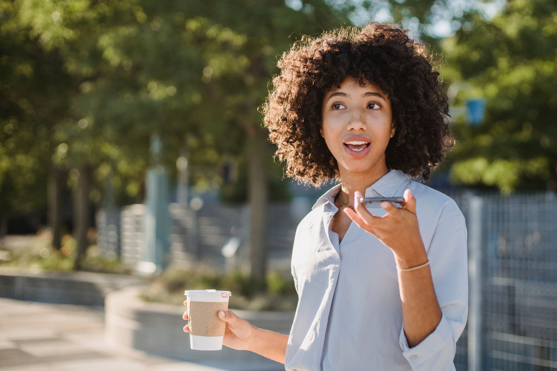 A woman walking outdoors speaking into a phone held near her mouth, a takeaway coffee cup in her other hand