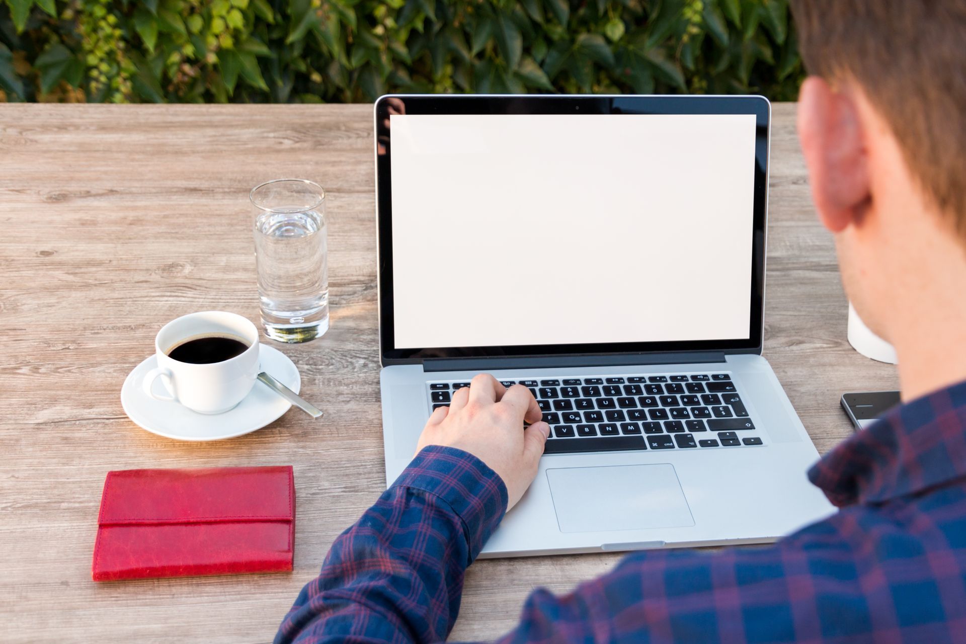 Seen over someone's shoulder, a laptop open on an outdoor wooden table beside an espresso cup, a glass of water and a red wallet