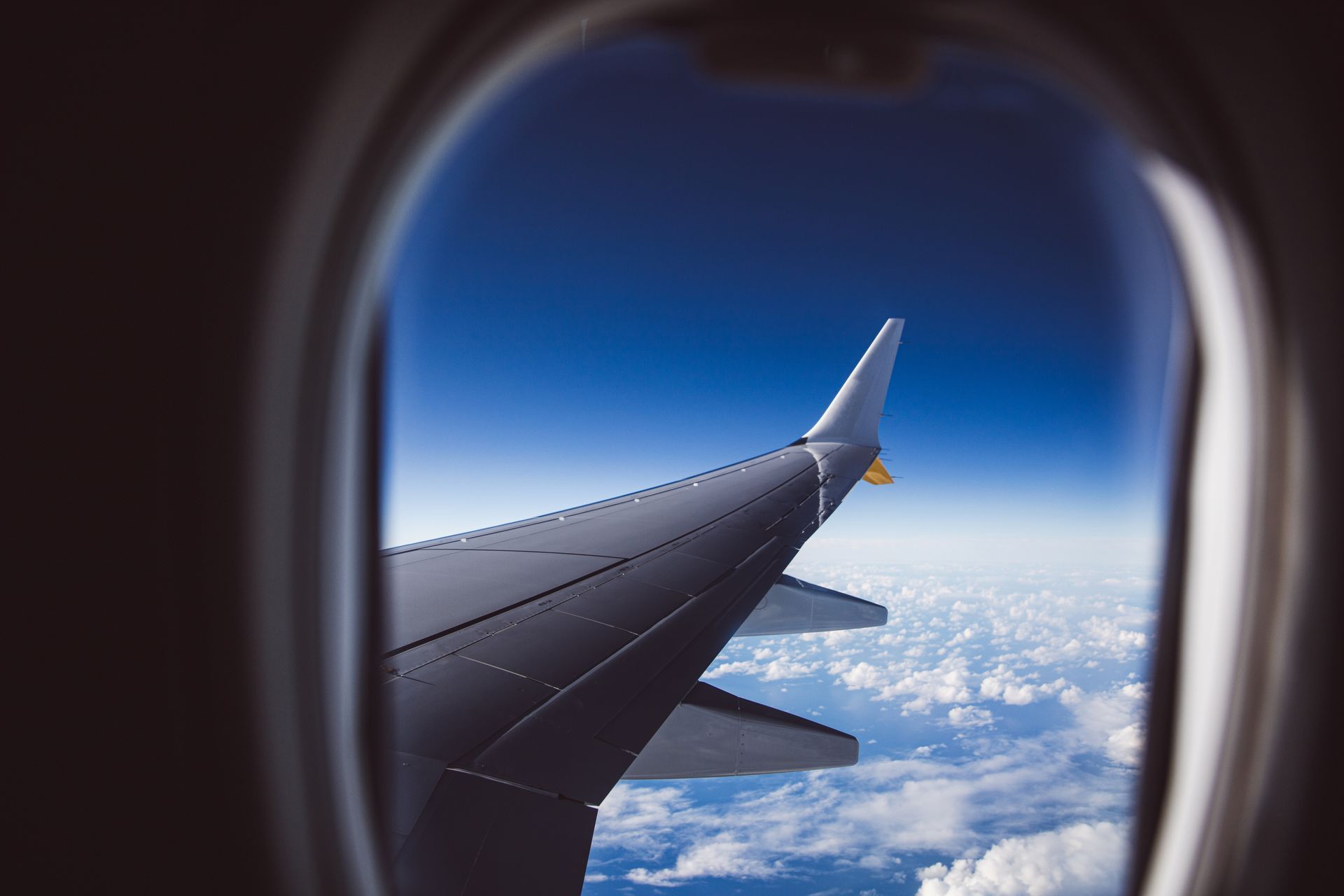 The wing of an aircraft seen through a cabin window, above a layer of cloud under a deep blue sky