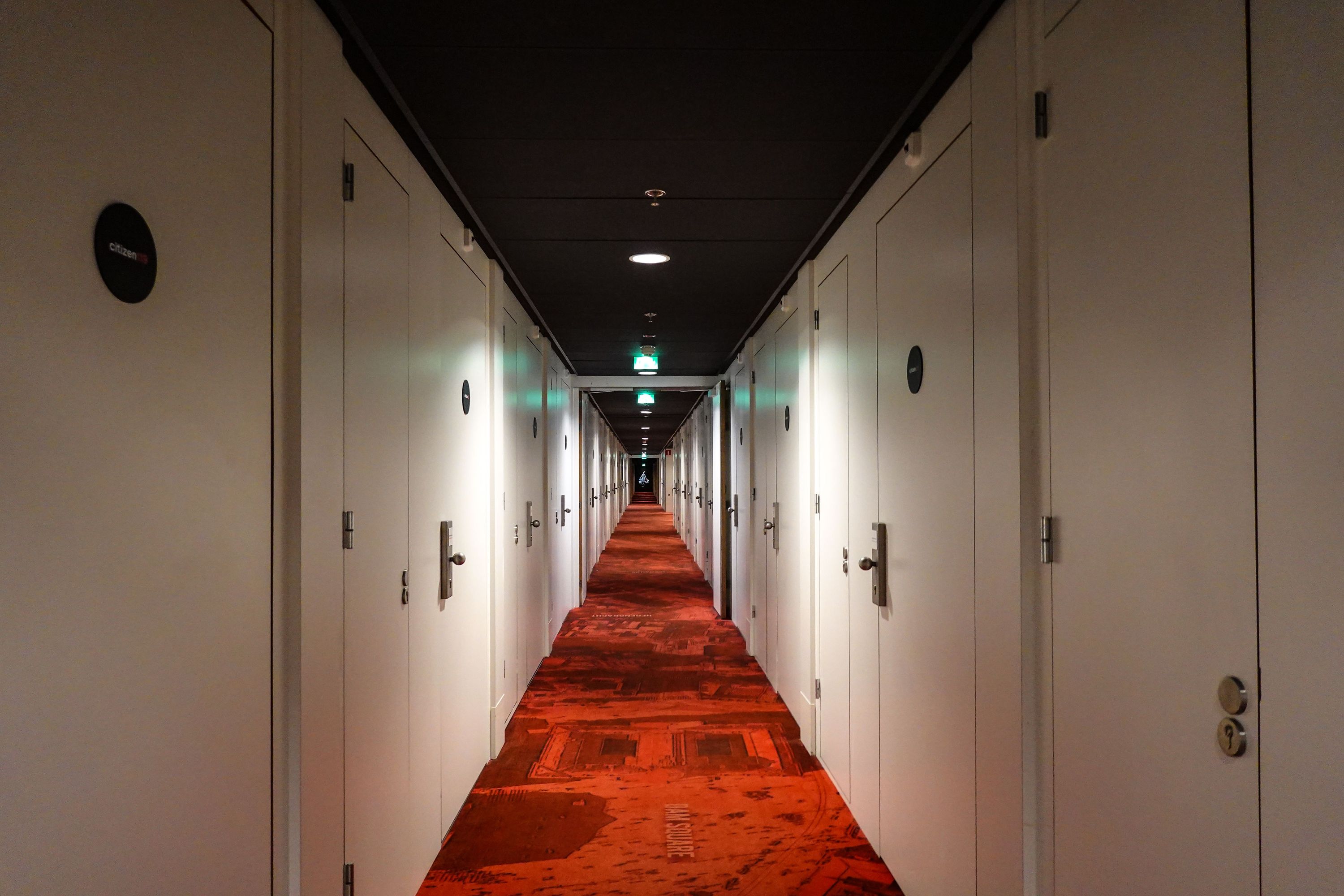 A long hotel corridor lined with identical white doors, red patterned carpet running to a lit stairway at the far end
