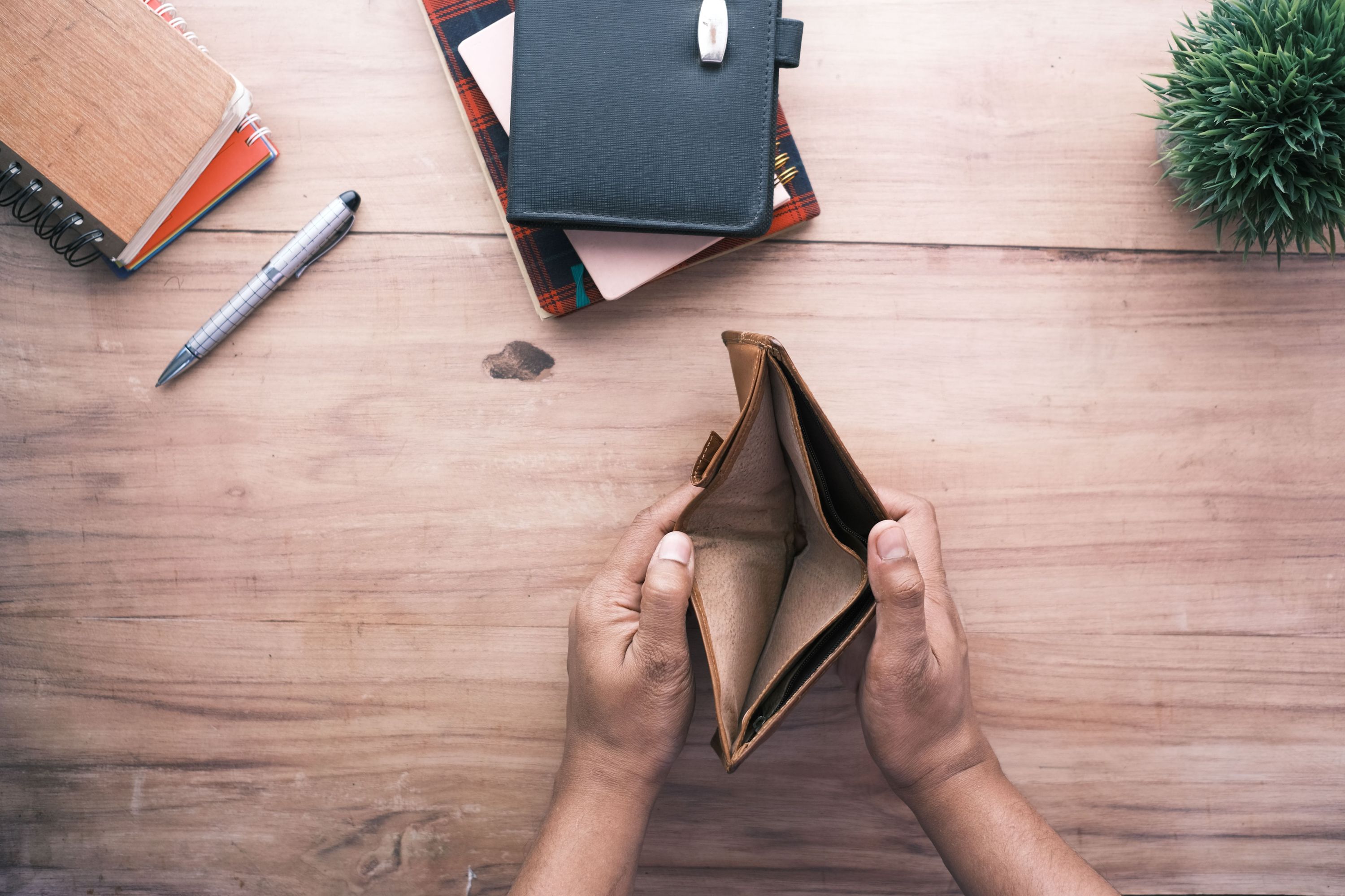 Two hands holding an open empty wallet above a wooden desk with notebooks, a pen and a small plant