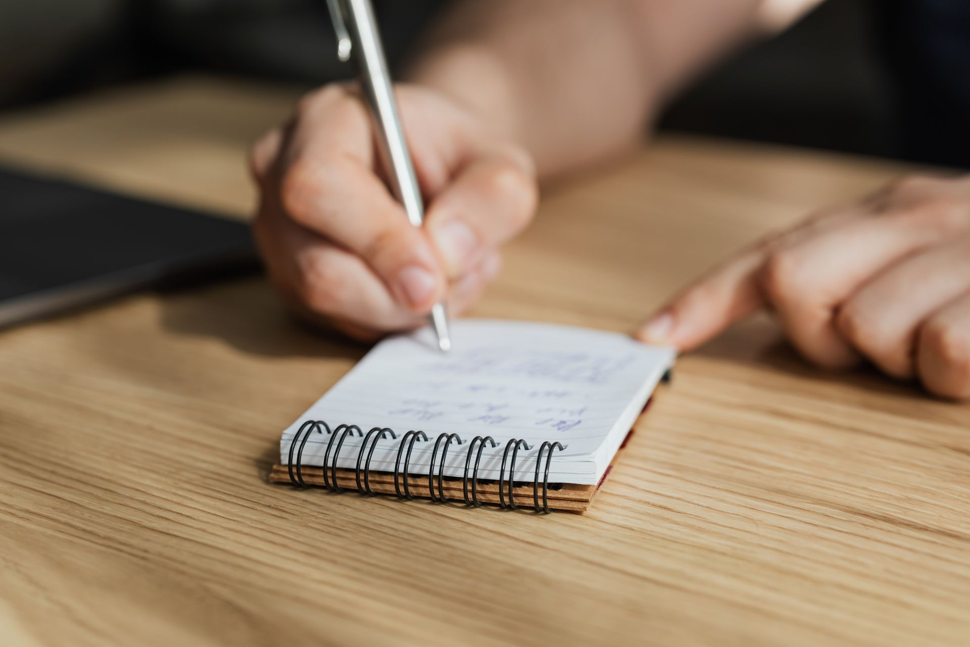 A close view of a hand writing a short line in a small spiral-bound notepad on a wooden table